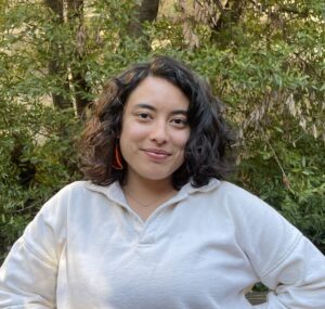 A headshot of Andrea smiling, she has short black shoulder length hair. She is wearing a white sweater with orange earrings. In the background there is a patch of tall bay laurel trees.