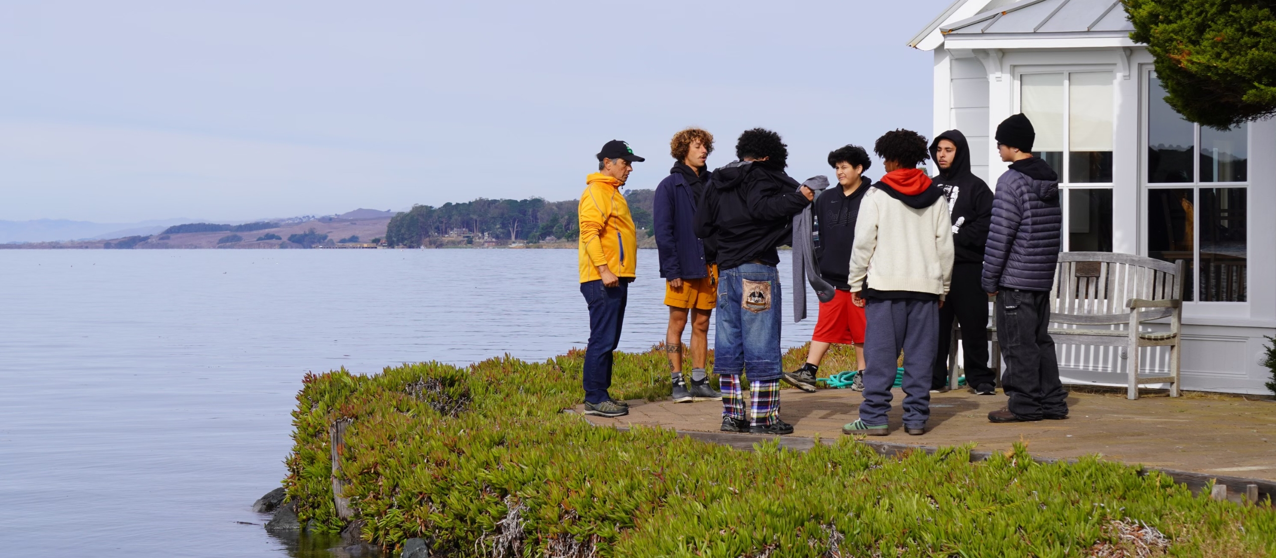 Youth gather in a circle along a shoreline in front of a white building.