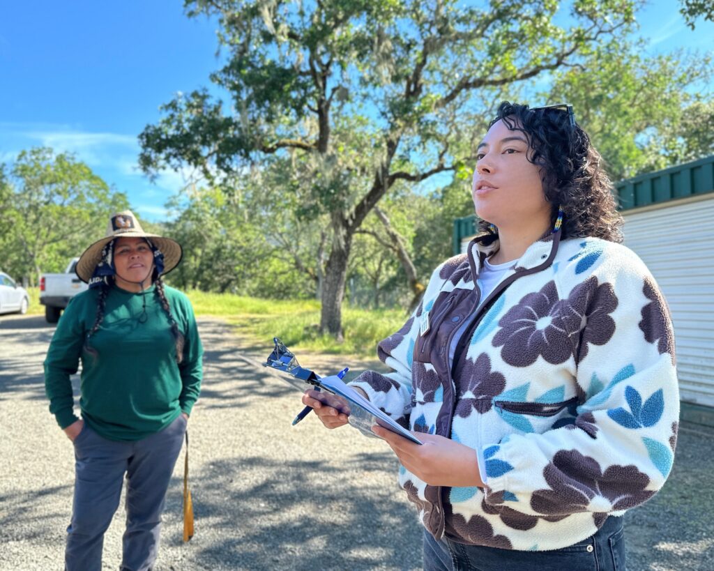 A woman speaks to a group while another woman looks on in the background.