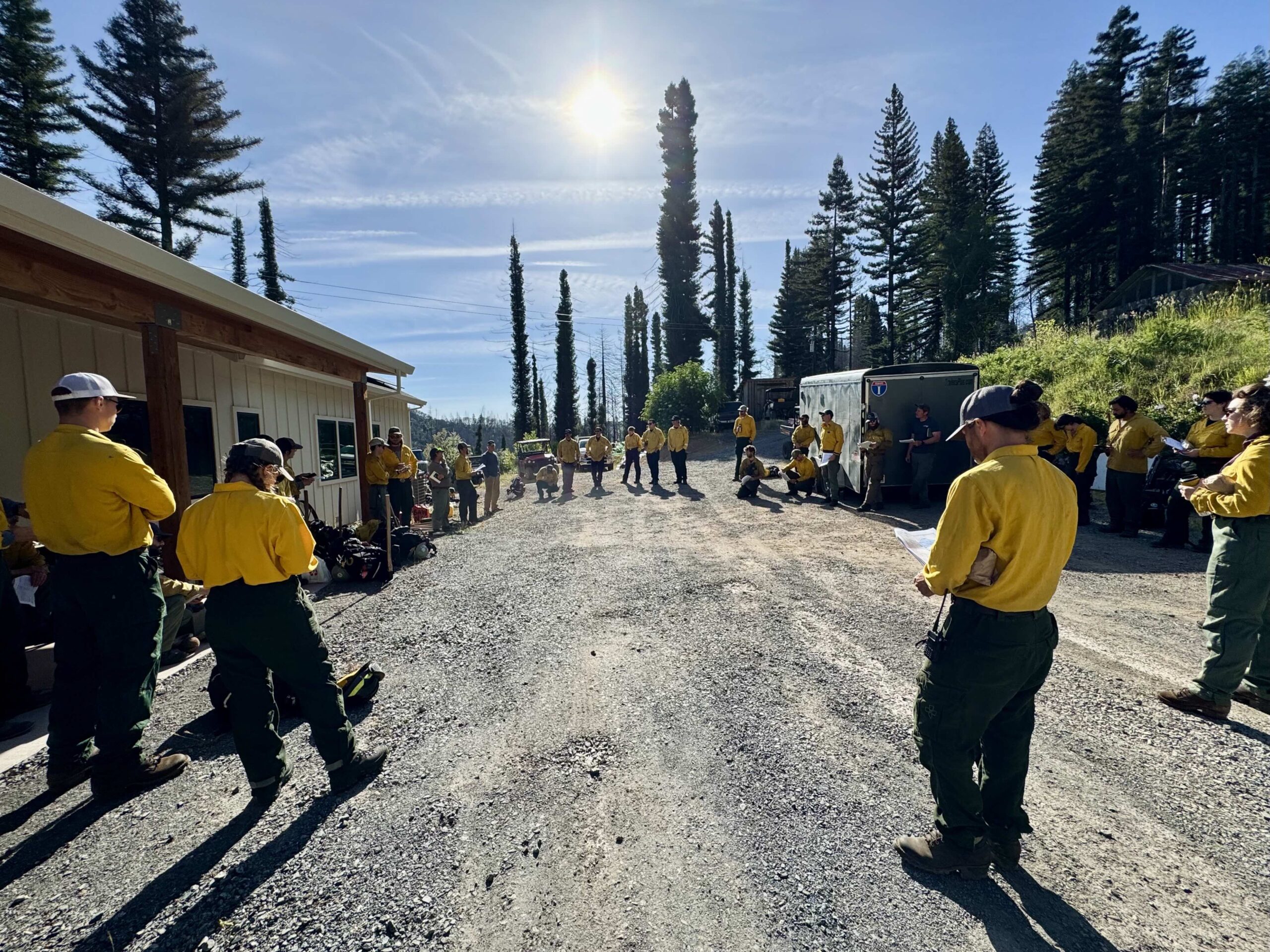 A group of workers in yellow shirts gather in a circle in the sun in a gravel driveway. 