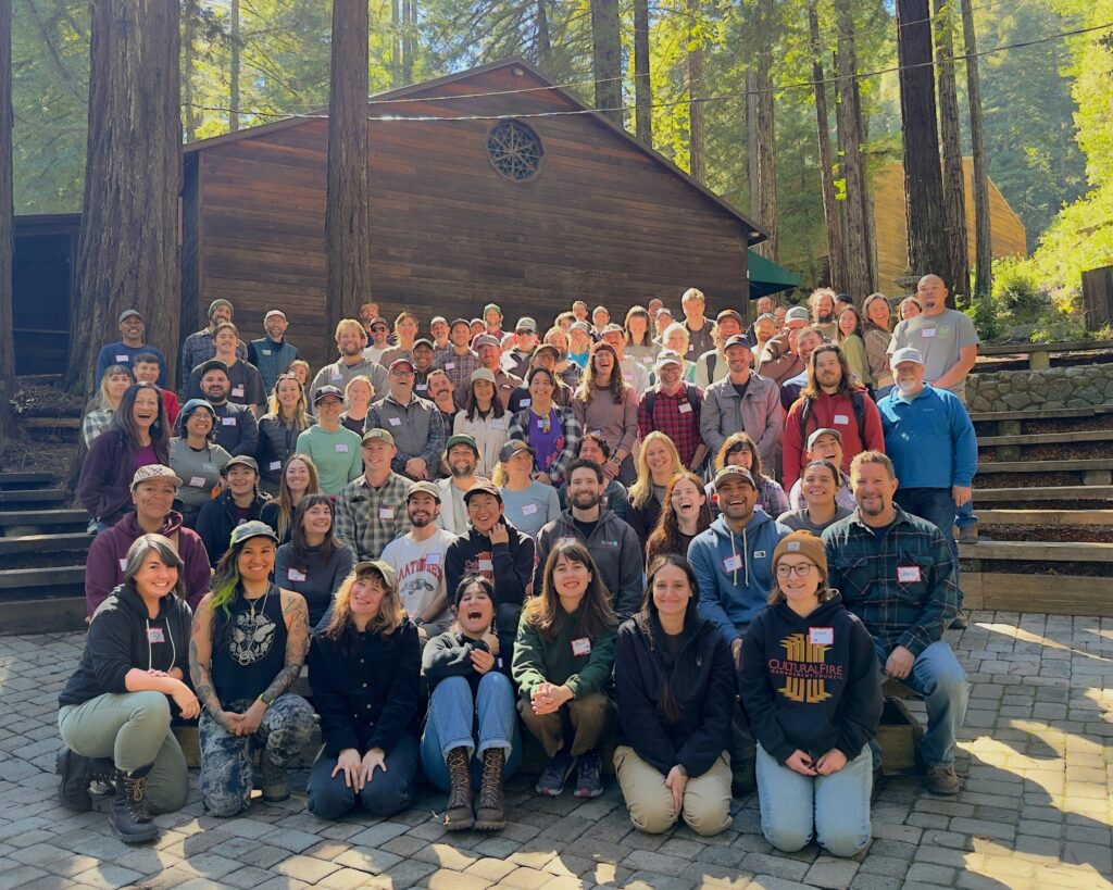 A group of more than 50 people of many ages, genders, and background sit in rows in an amphitheater posing for the camera in a redwood grove