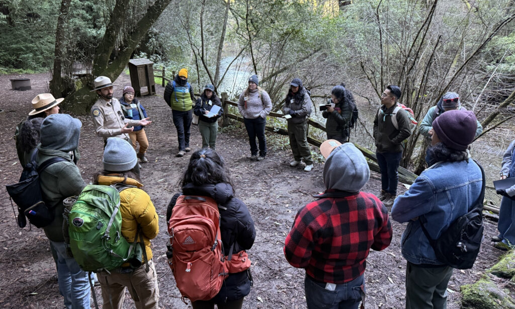 A group of people of mixed ages gather in a circle beside a stream and listen to a parks worker talk.