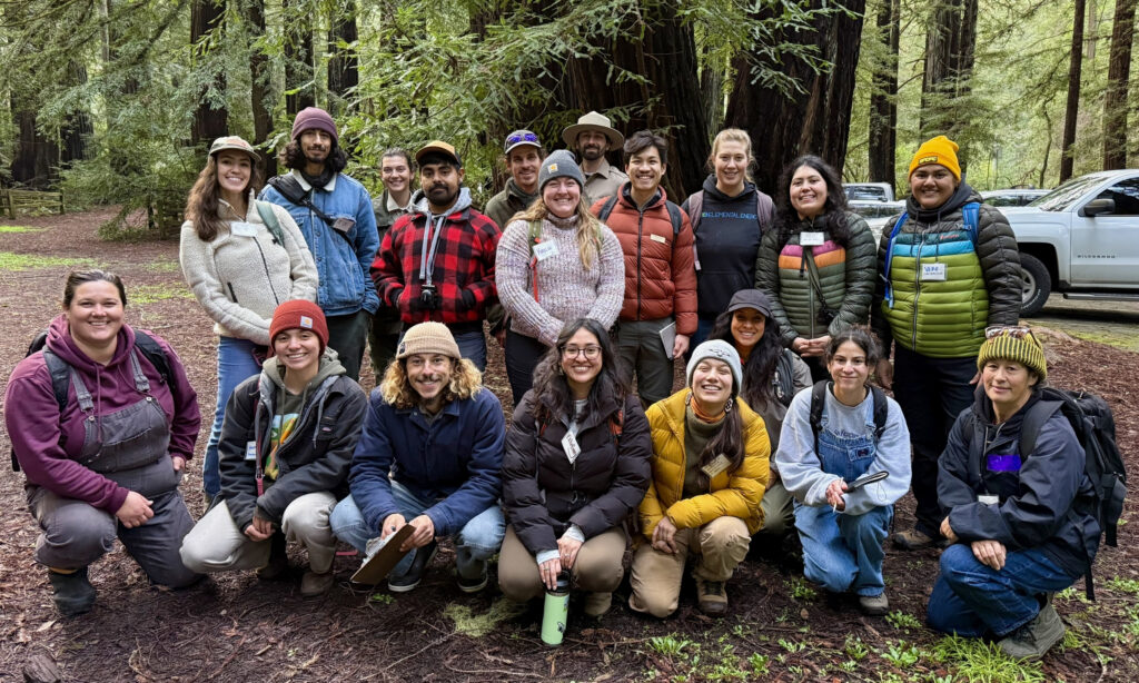 A group of people of mixed ages and backgrounds in coats and hats standing in a redwood forest smile and face the camera.