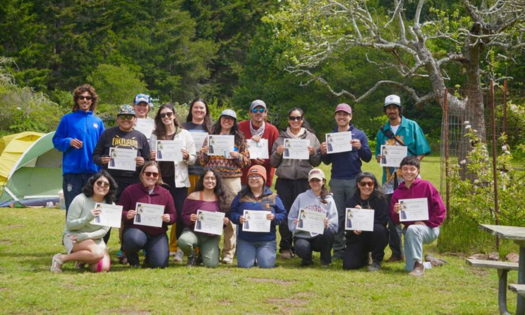 A group of people of mixed ages and backgrounds stands in two rows facing the camera smiling holding up certificates. 