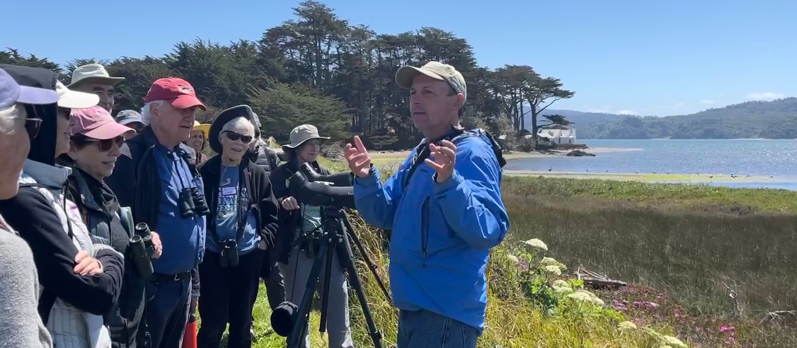 A man in a blue jacket giving a talk along the shore of a wetlands.