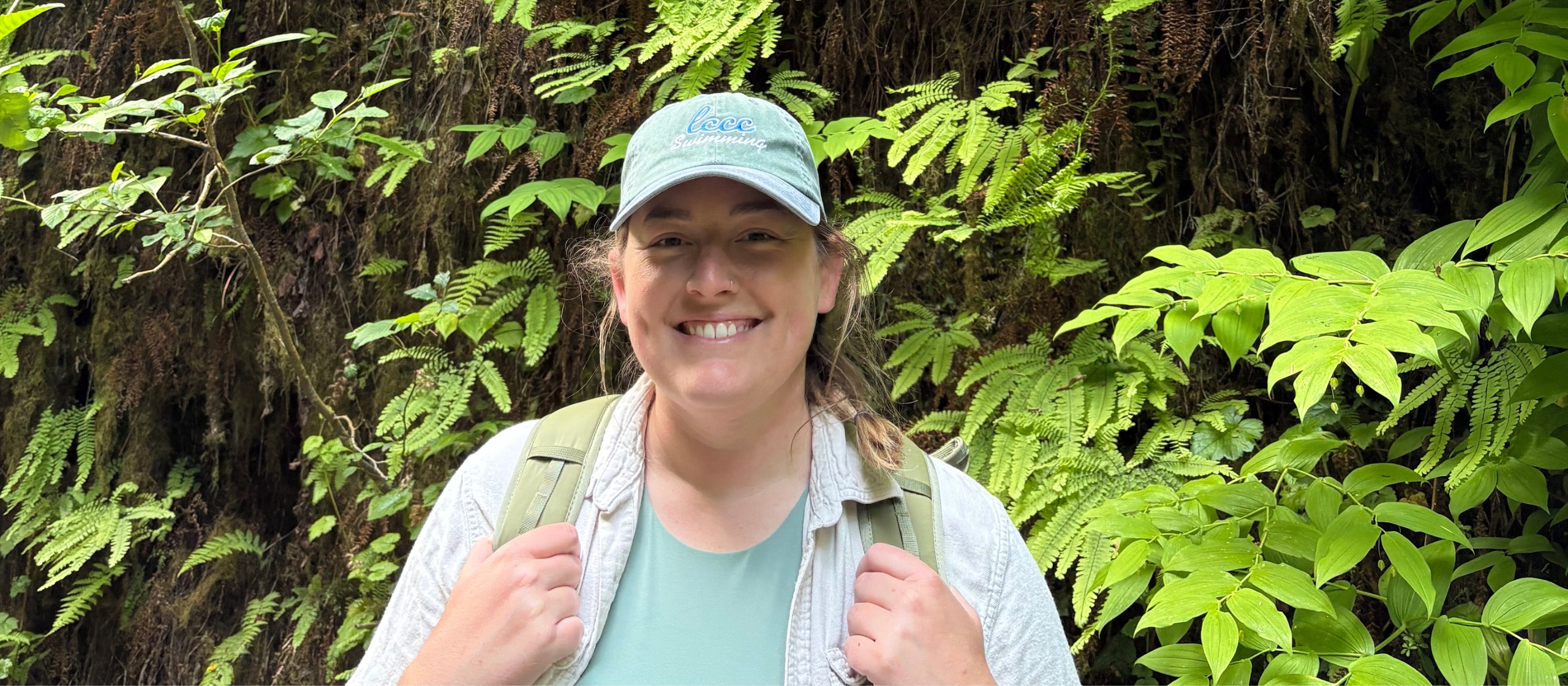 A woman in a hat faces the camera smiling, behind her is are bright green ferns.
