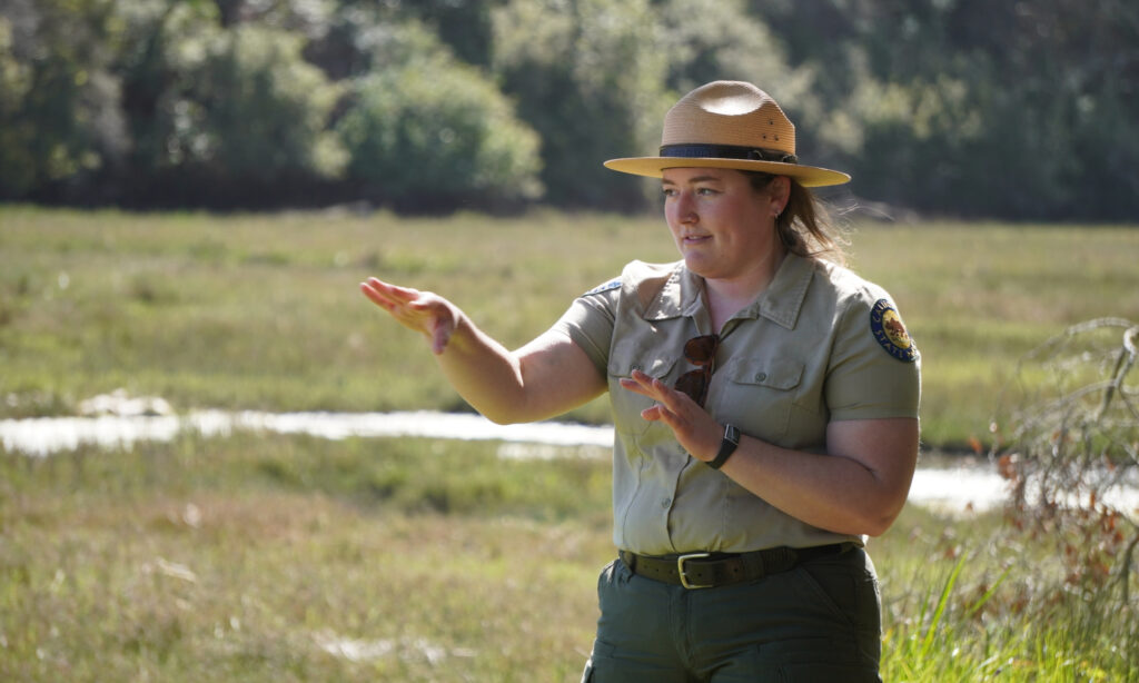 A State Parks person in uniform giving a talk with a wetland behind her. 