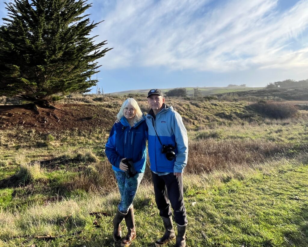 A man and woman stand side by side, arms around each other with a grassland and blue sky with clouds behind them