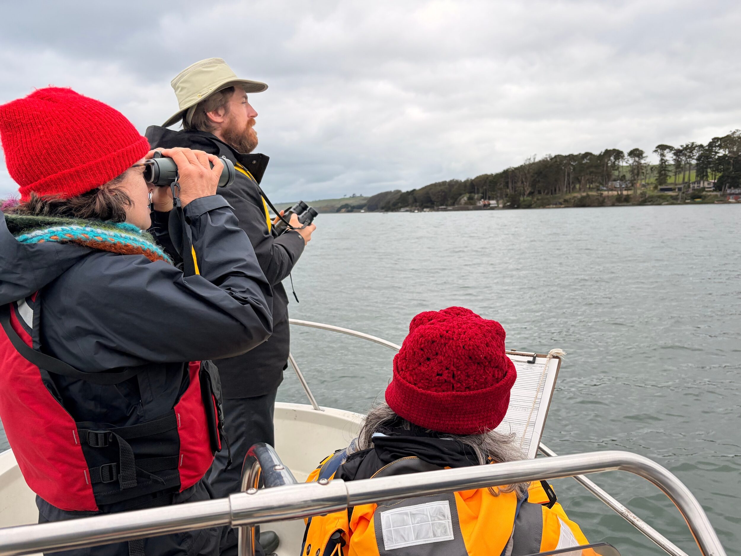 A group of three people watching birds on a boat in a bay with binoculars and a clipboard.