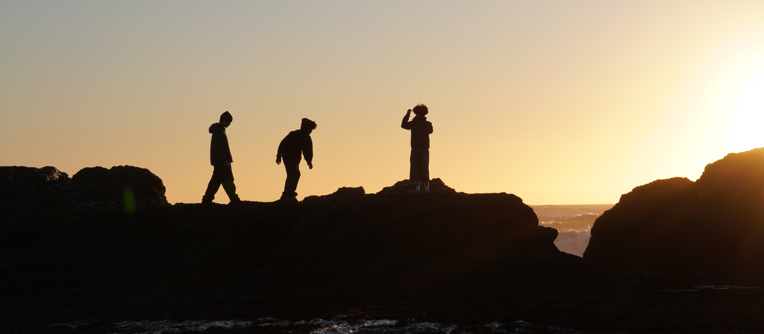 Three silhouettes stand on a rocky outcropping by the ocean with the sunset behind them.