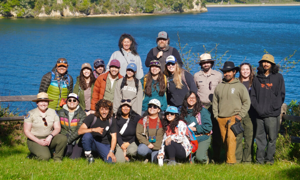 A group of 20 people in three rows in from of a coastal landscape on a sunny day.