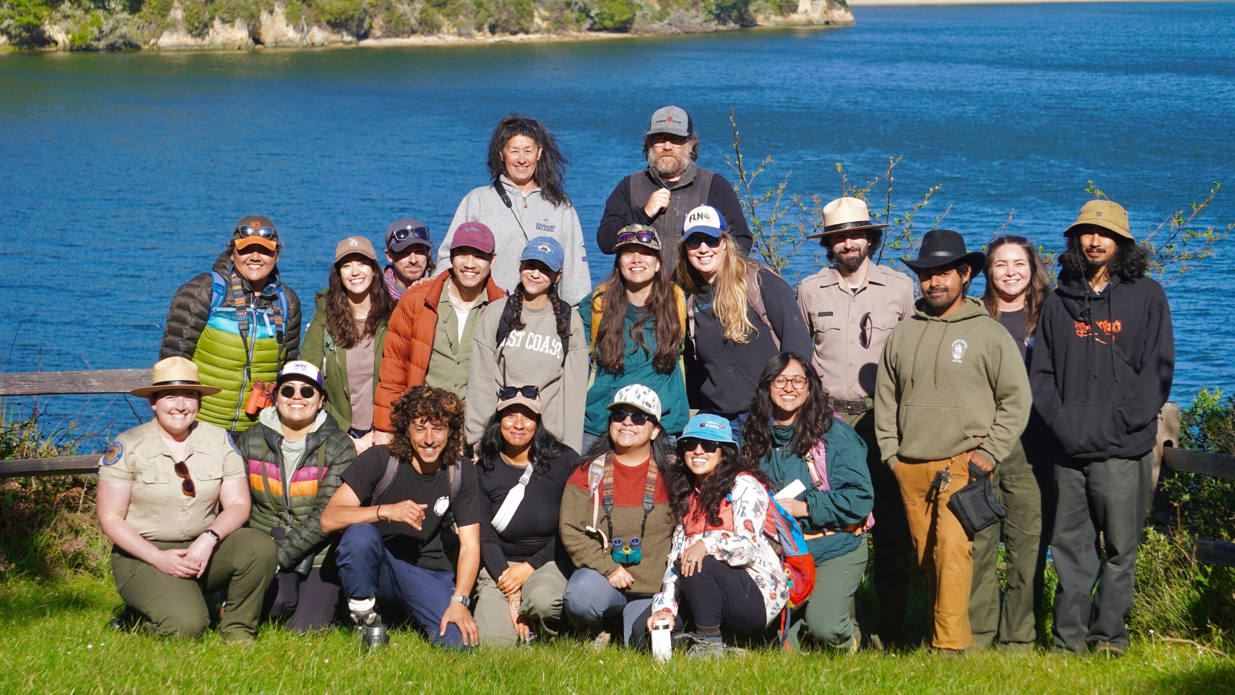 A group of 20 people in three rows in from of a coastal landscape on a sunny day.