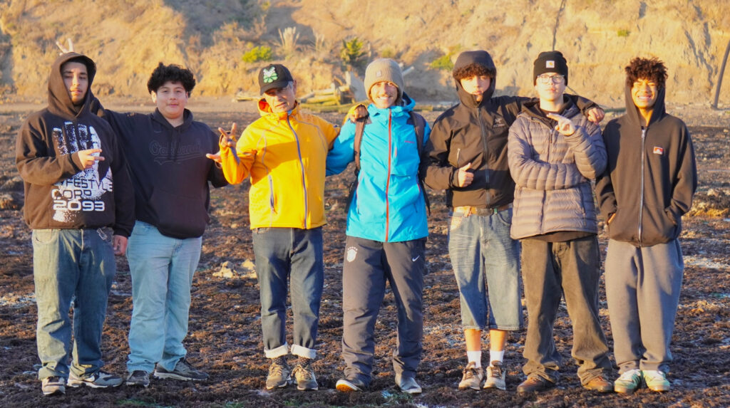 A group of youth with an adult standing in a line in rain gear in a seaside location.