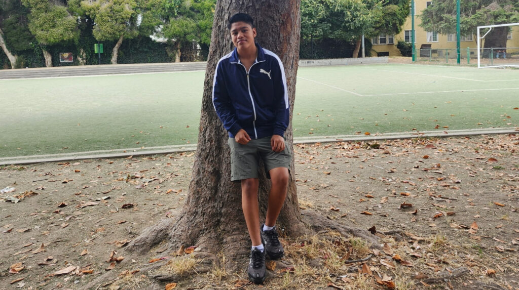 A teenager in a black track jacket standing casually against a tree with a soccer field behind him.