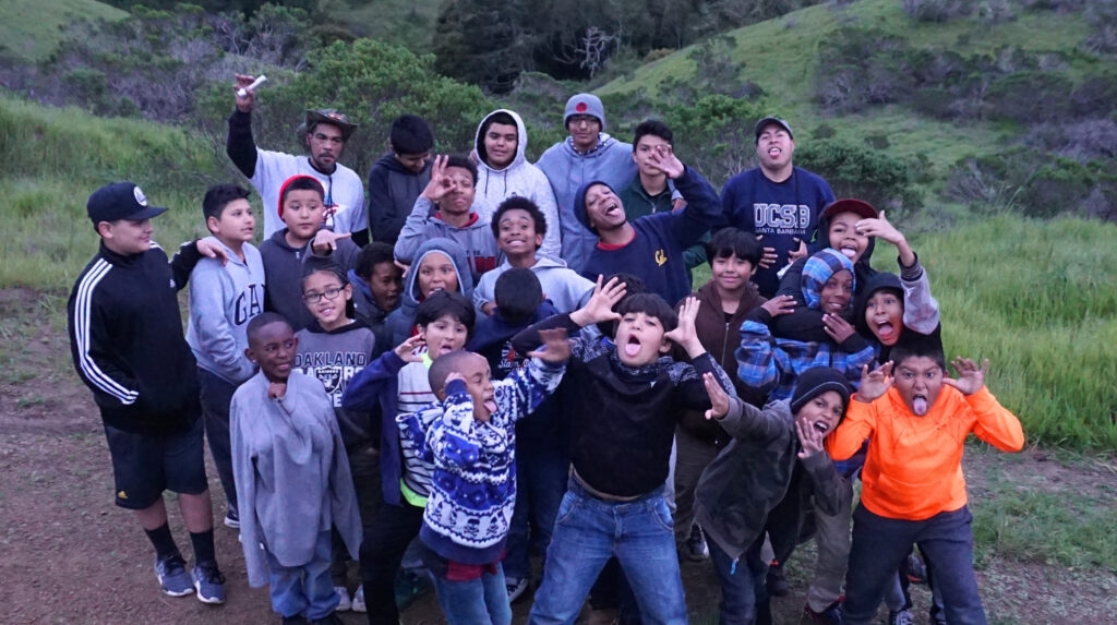 A group of mixed ages boys, teenagers, and men making funny faces on a forest trail with trees behind them.
