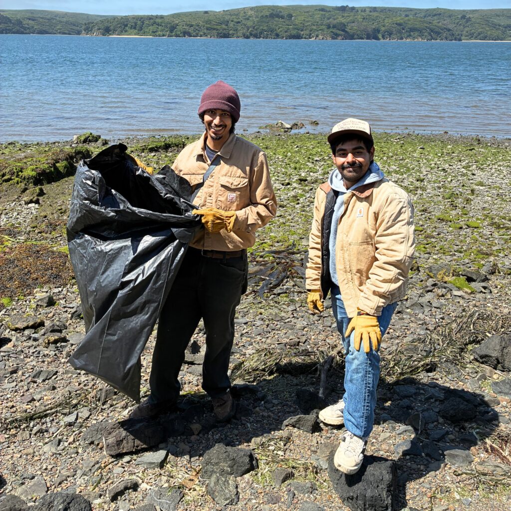 Two young men of medium skin tone in work clothes are working on a shoreline with a trashbag doing a coastal clean up.