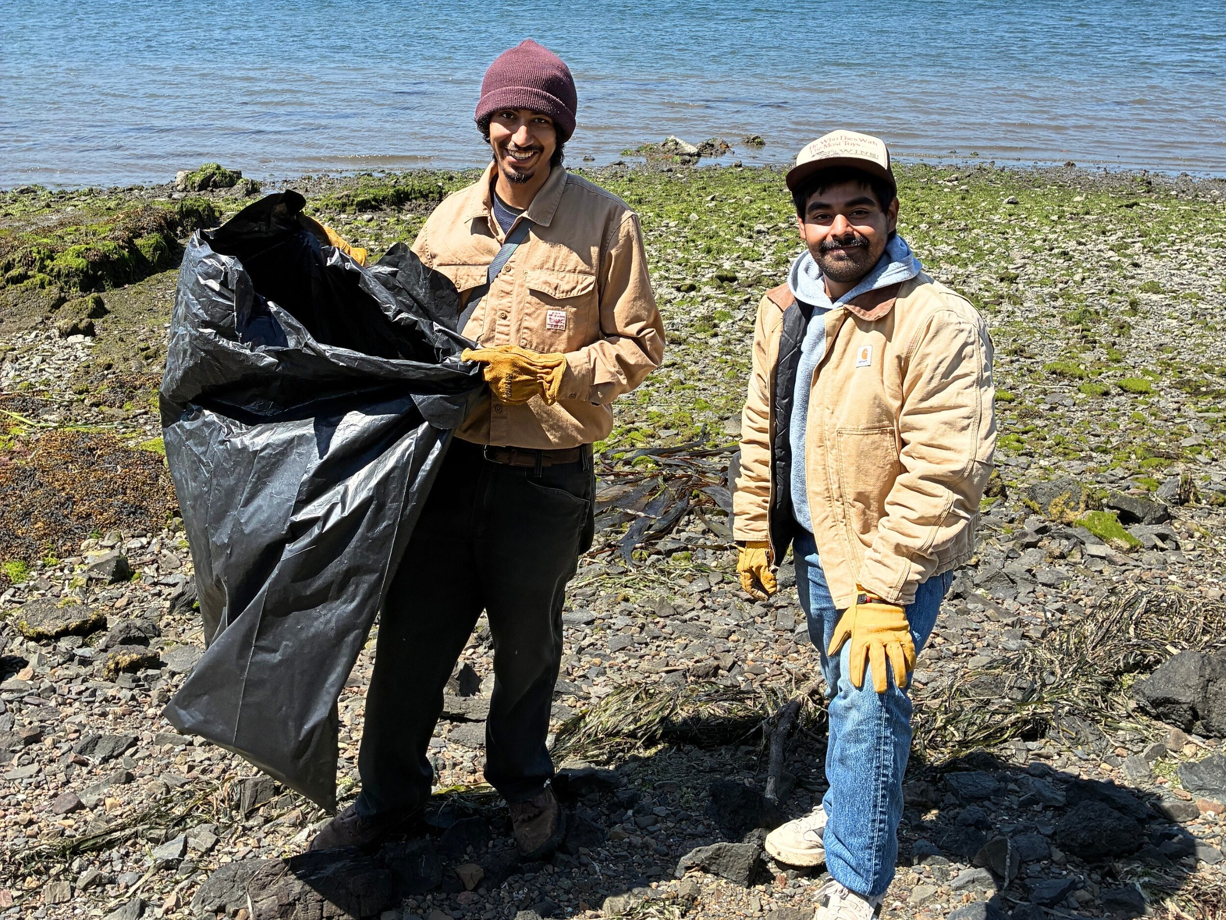Two young men of medium skin tone in work clothes are working on a shoreline with a trashbag doing a coastal clean up.