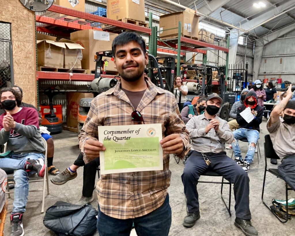 A young man with medium skin tone holds up a certificate smiling with people seated behind him applauding.