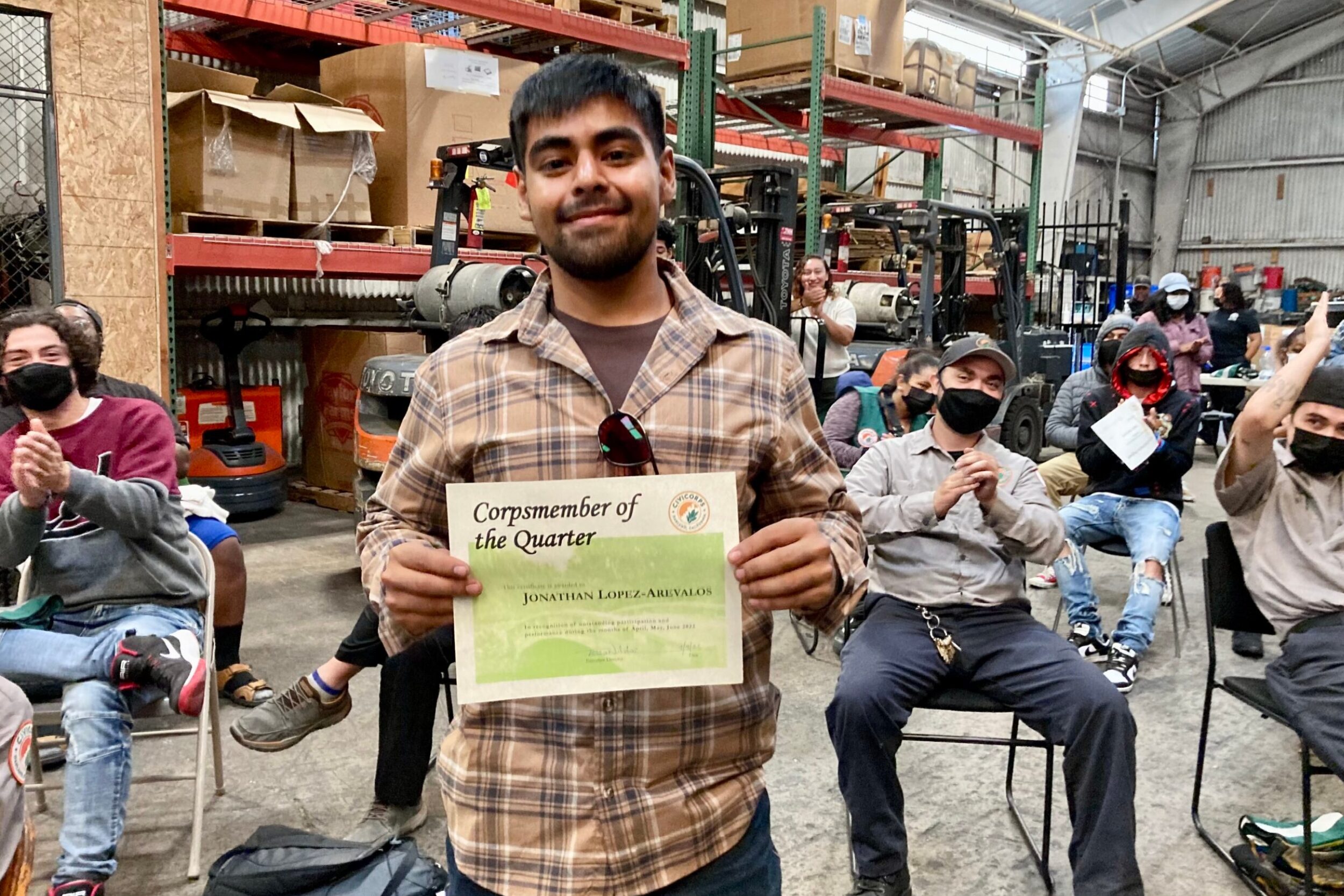A young man with medium skin tone holds up a certificate smiling with people seated behind him applauding.