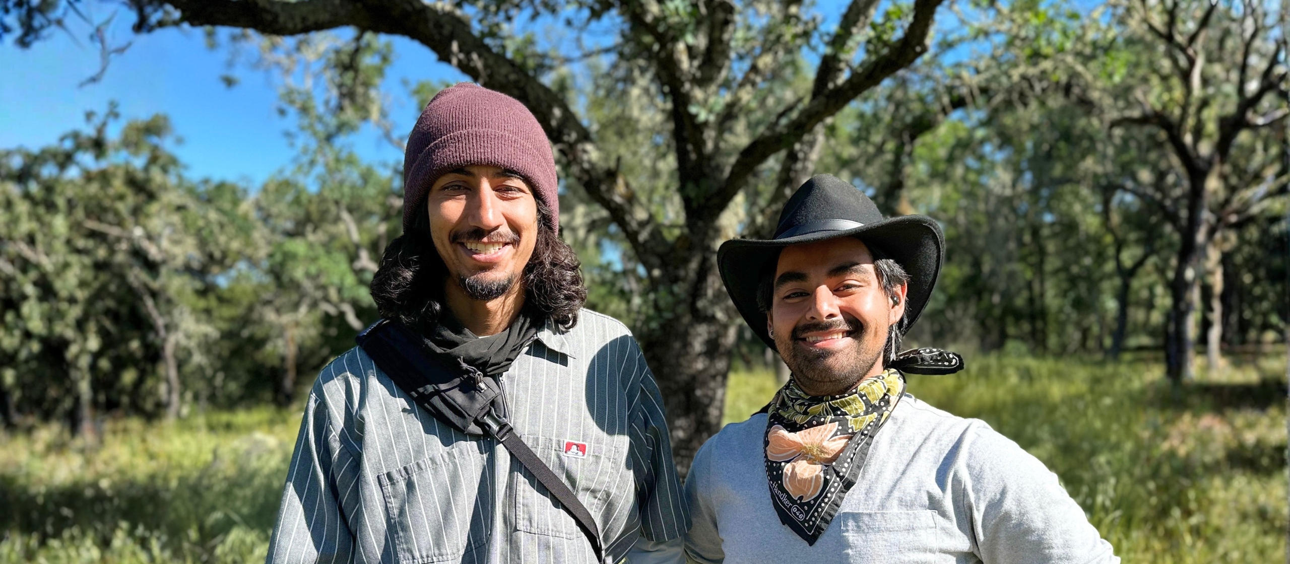 Two young men of medium skin tone smile in front of a landscape of oak trees and green grass.
