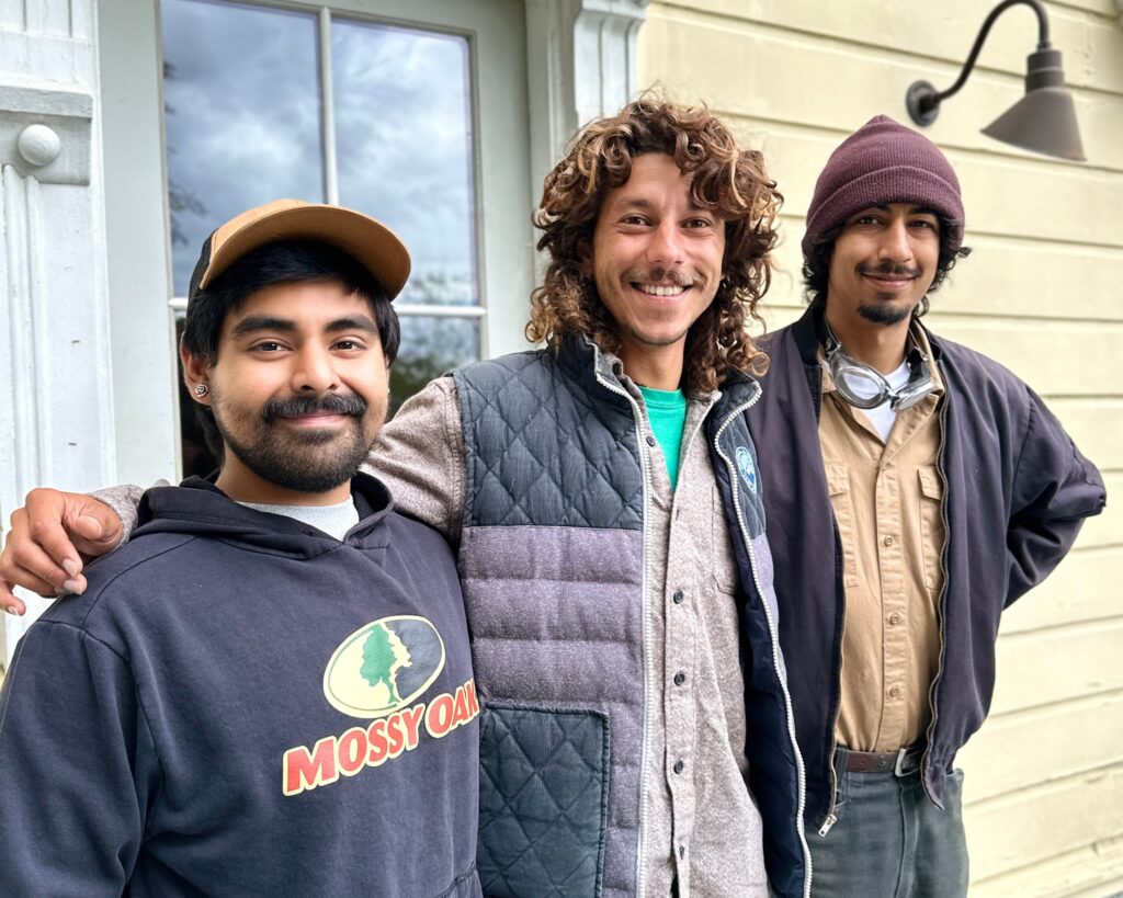 Three young men face the camera smiling in front of the door of a house. 