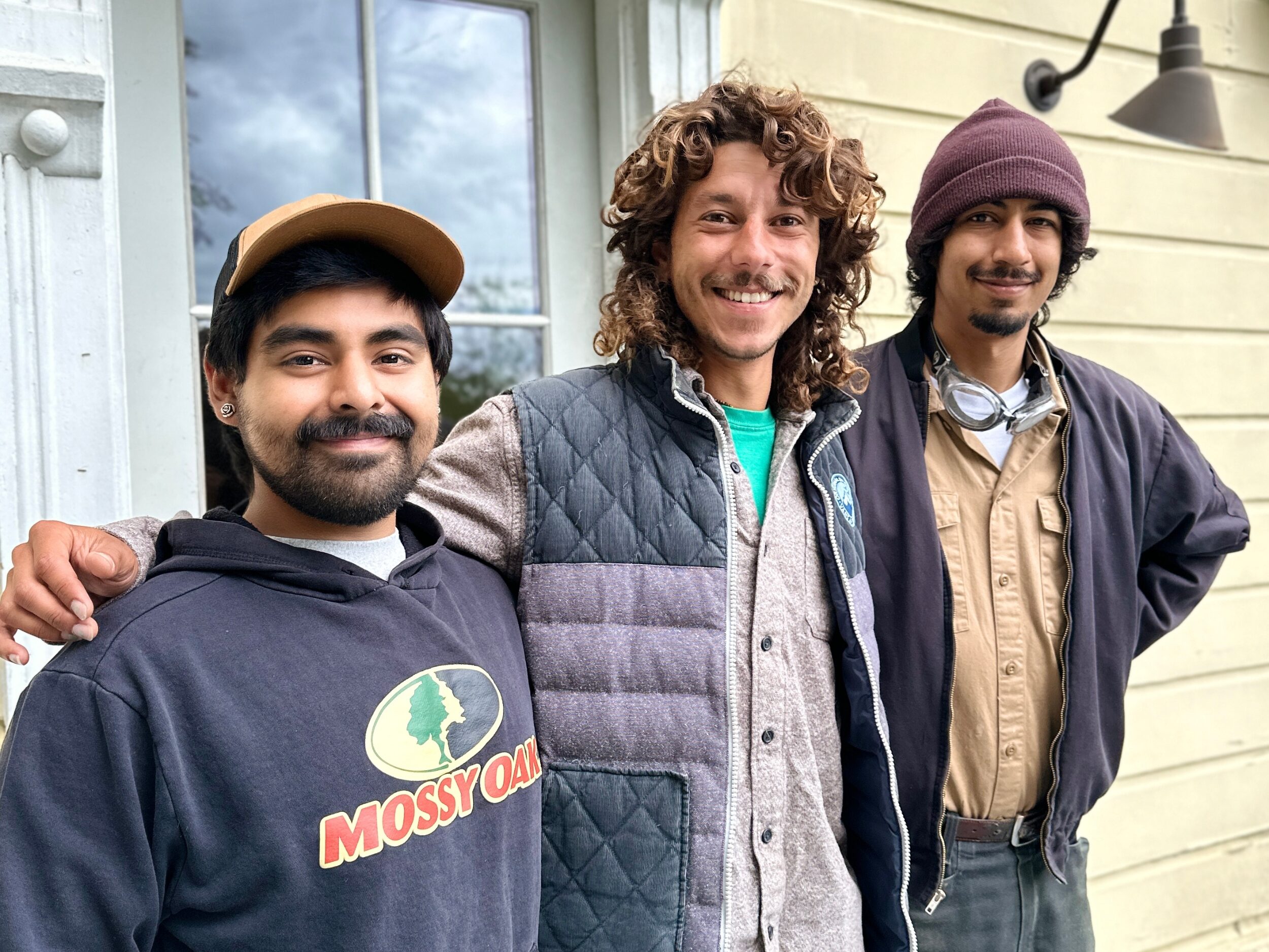 Three young men face the camera smiling in front of the door of a house. 