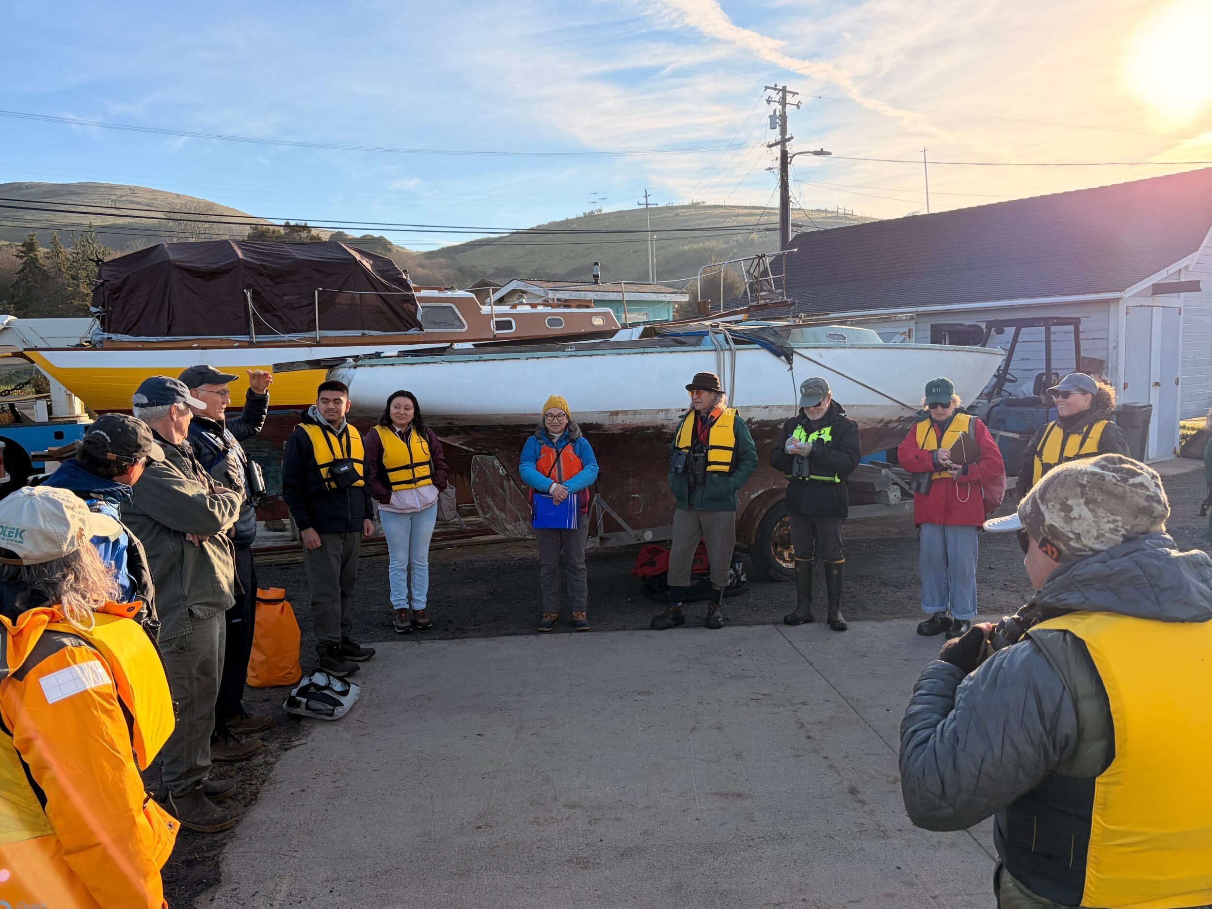 A group of 20 people of mixed ages and gender with lifejackets and binoculars gathers in a circle in the early morning sun beside the water.