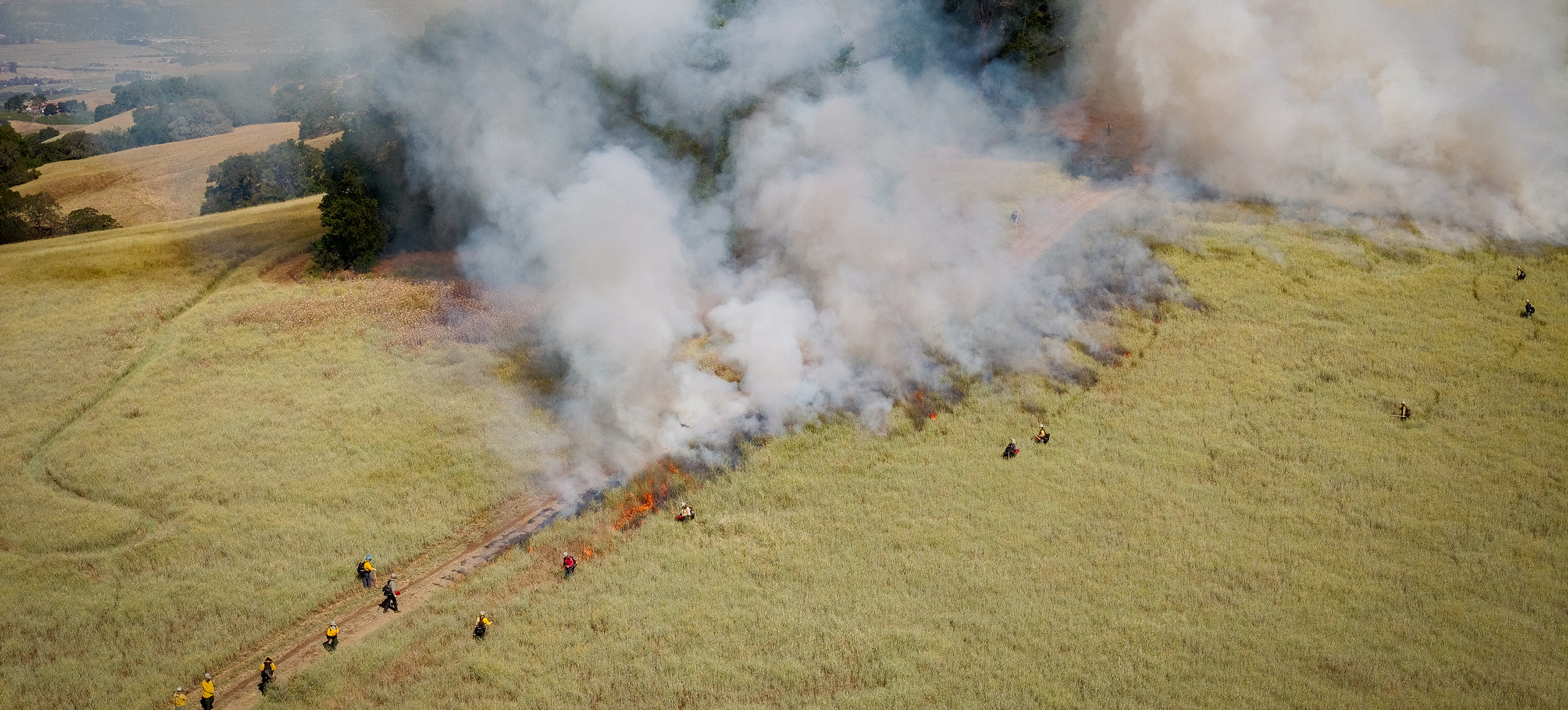 An aerial view of prescribed fire in a dry grassland with smoke rising up in the air.