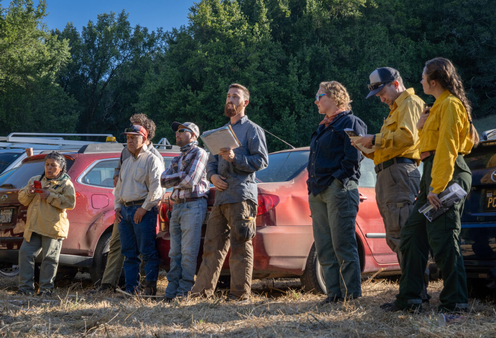 A man addressing a groupof outdoor workers some of them in safety gear outdoors in a parking lot.