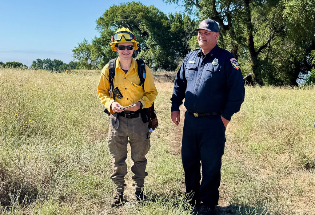 A man in yellow safety gear and a hard hat stands laughing with a uniformed emergency worker in blue in a field of dry grass.