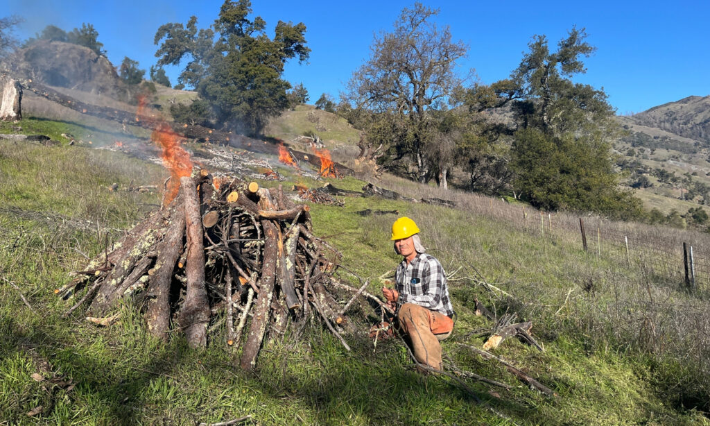A man in a yellow hard hat and safety kneels next to a pile of wood on fire.