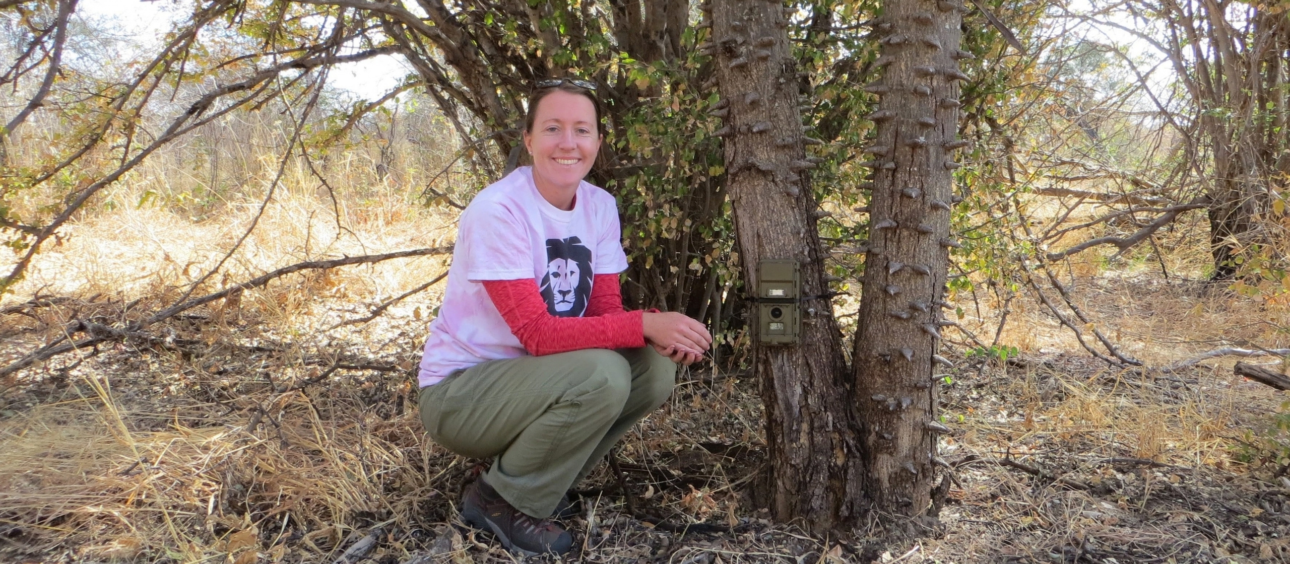 A women in a pink t-shirt crouches next to a tree setting up a trail camera.